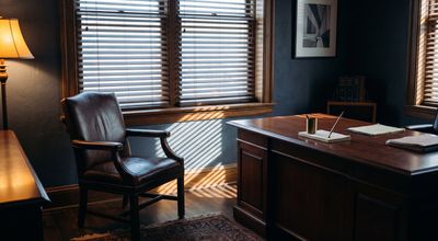 A sparse law office with an empty leather chair at a polished desk, blinds striping the afternoon light across it.
