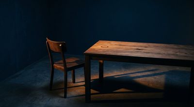 An empty chair pulled back from a plain table, recently vacated, amber light raking low across the surface.